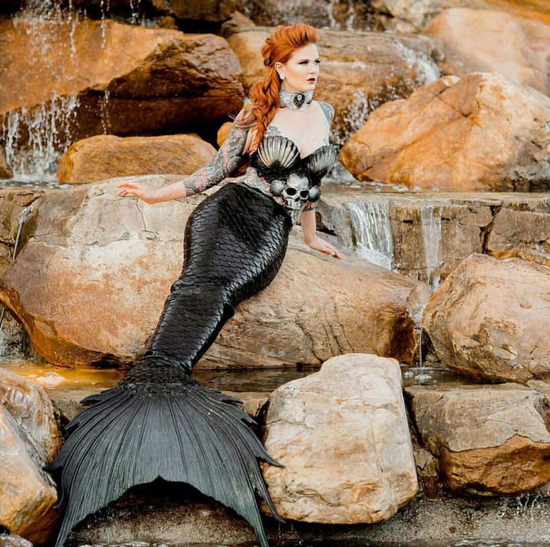 美人魚 桌布 壁紙 — 一位紅髮美人魚，擁有黑色鱗片尾巴和骷髏主題上衣，在瀑布旁的岩石上優雅地擺姿勢。
