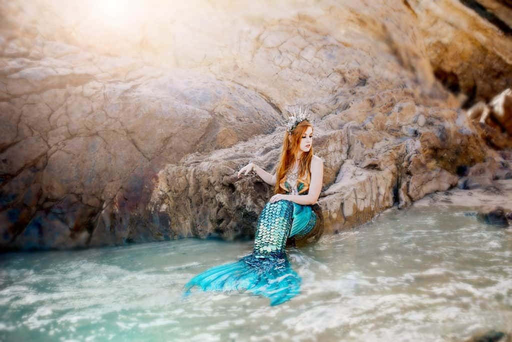 Mermaid Real Life 桌布 壁紙 — 一位紅髮美人魚閃爍著藍色尾巴，優雅地坐在岩石海岸上，金色陽光灑滿整個場景。