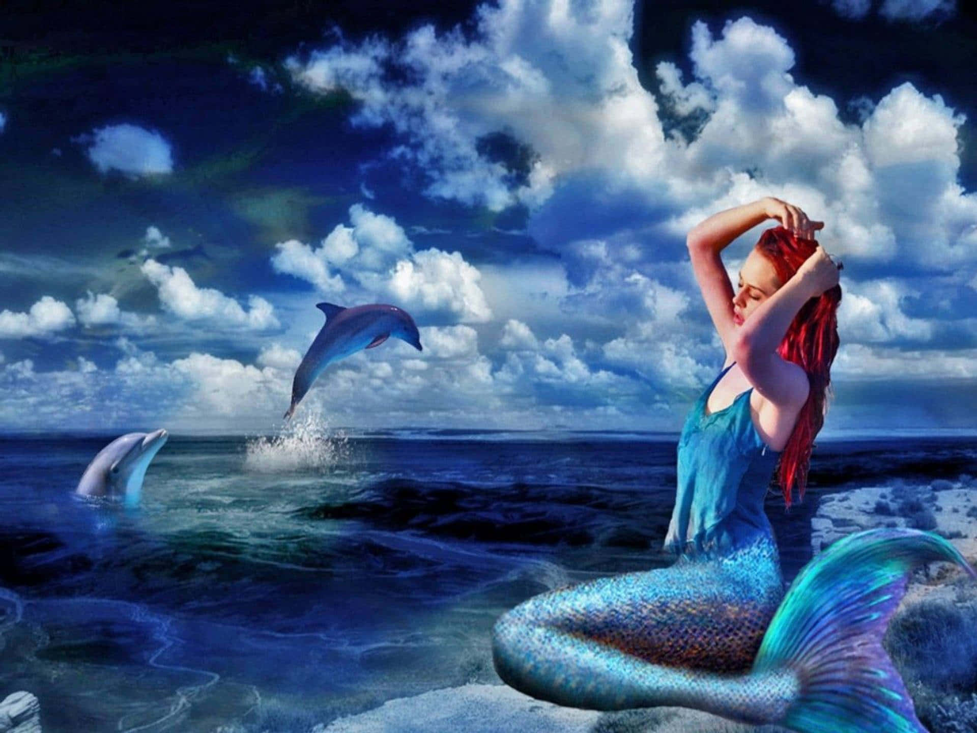 dolphins 桌布 壁紙 — 一位紅髮藍尾的美人魚坐在岩石海岸上，兩隻海豚在戲劇性的多雲天空下在海洋中跳躍。