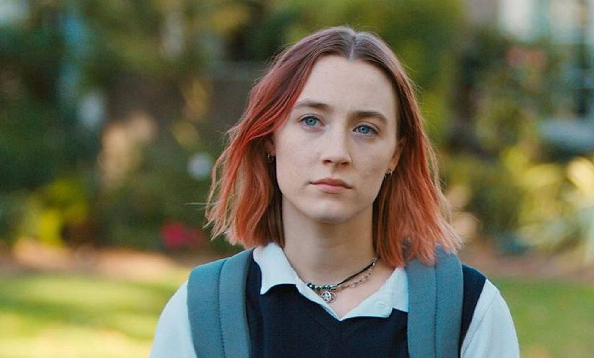 Saoirse Ronan 桌布 壁紙 — 一位紅髮藍眼的年輕女性背著背包，在陽光普照的戶外環境中直視鏡頭。