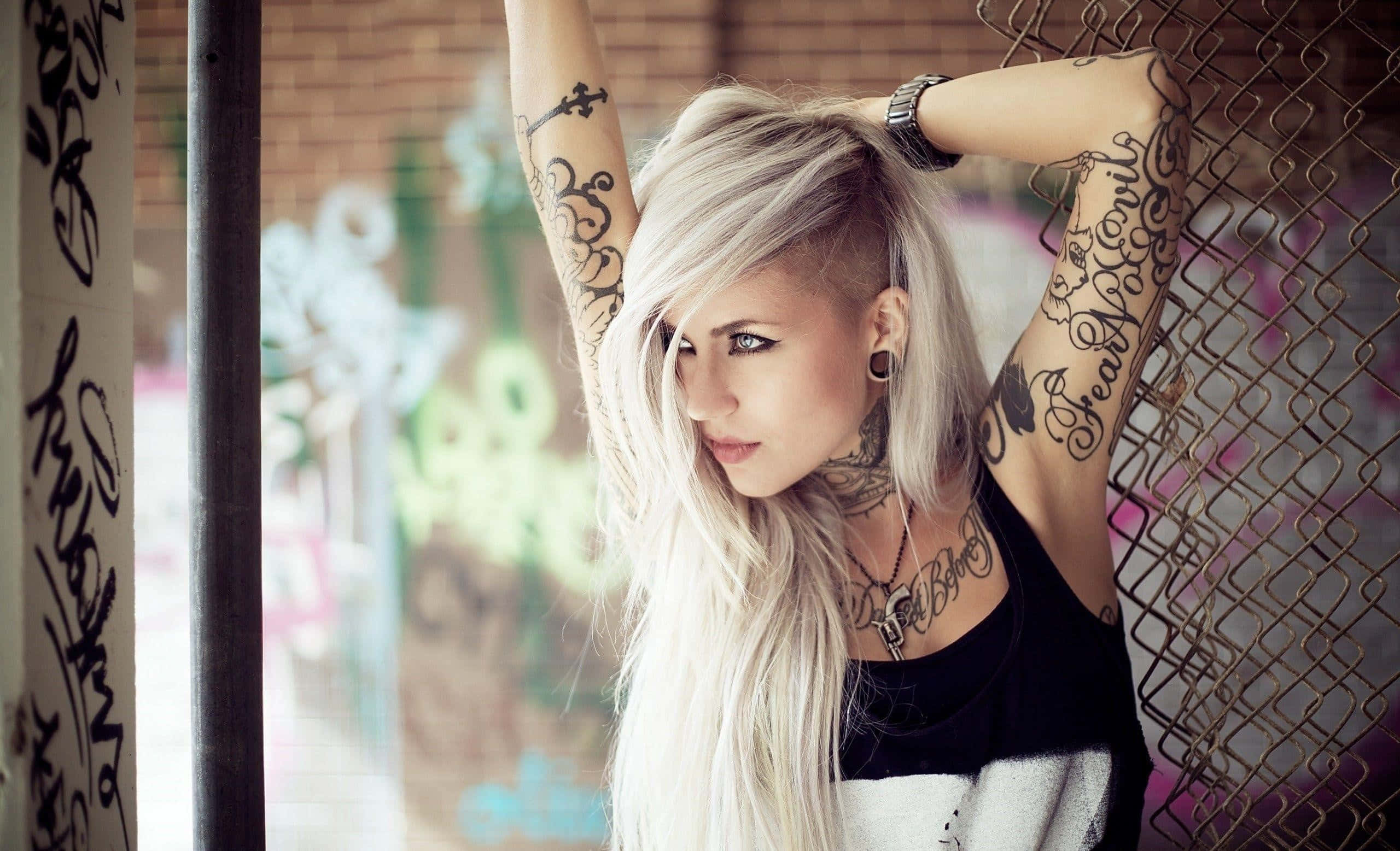 Tattoo 桌布 壁紙 — 一位紋身、鉑金色頭髮的女性靠在一個被塗鴉覆蓋的城市小巷中的鐵絲網圍欄上。