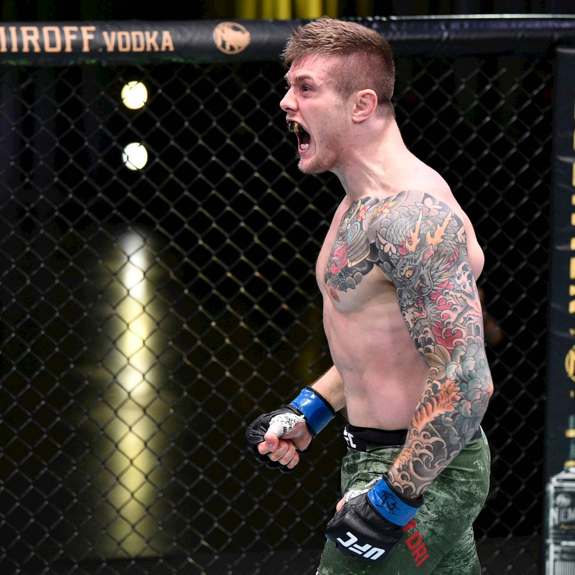 UFC 桌布 壁紙 — 一位紋身MMA拳手在籠內咆哮勝利，周圍有明亮燈光和贊助商標誌。