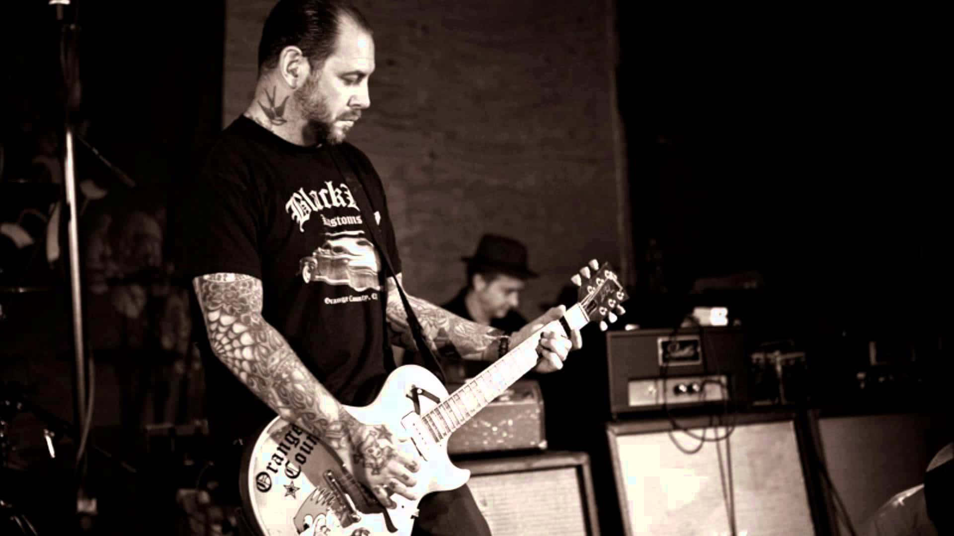 guitar player 桌布 壁紙 — 一位紋身吉他手在燈光昏暗的舞台上彈奏白色電吉他，背景有擴音器和鼓手。