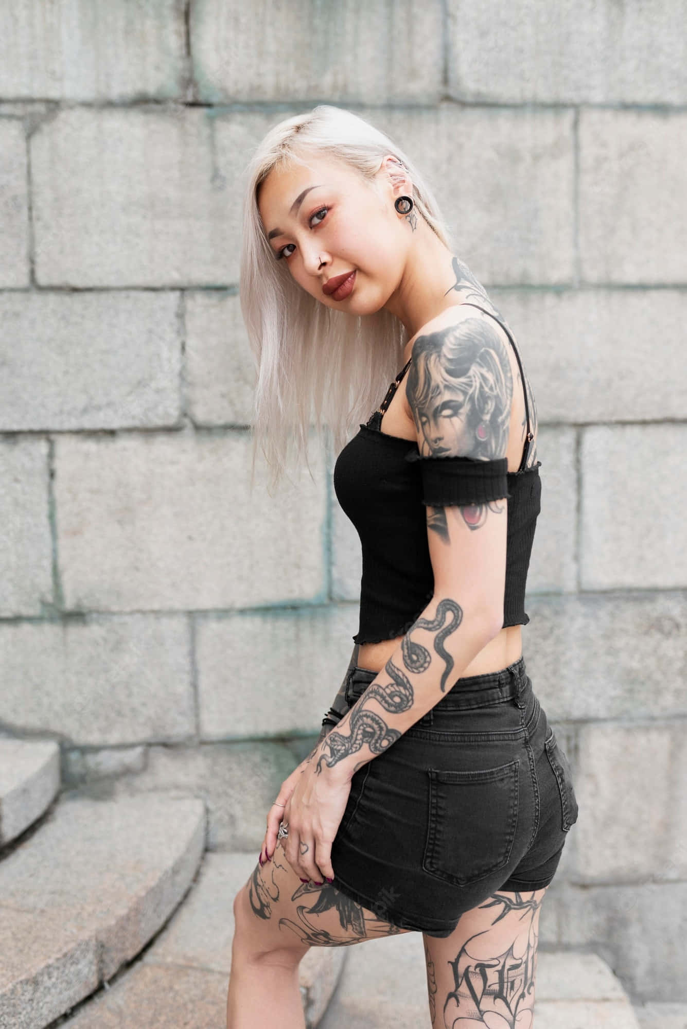 Tattoo Girl 桌布 壁紙 — 一位紋身女子，一頭鉑金色頭髮，身穿黑色短上衣和短褲，自信地靠在灰色石牆上擺姿勢。