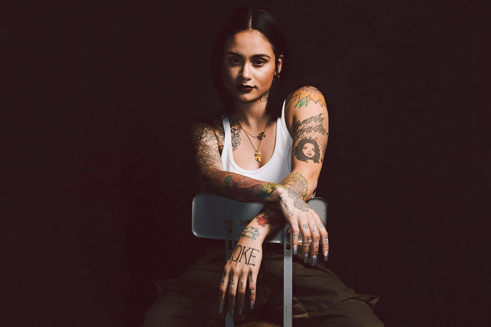 kehlani 桌布 壁紙 — 一位紋身女子，深色頭髮，自信地坐在黑色背景前的椅子上，雙臂交叉，目光直視。