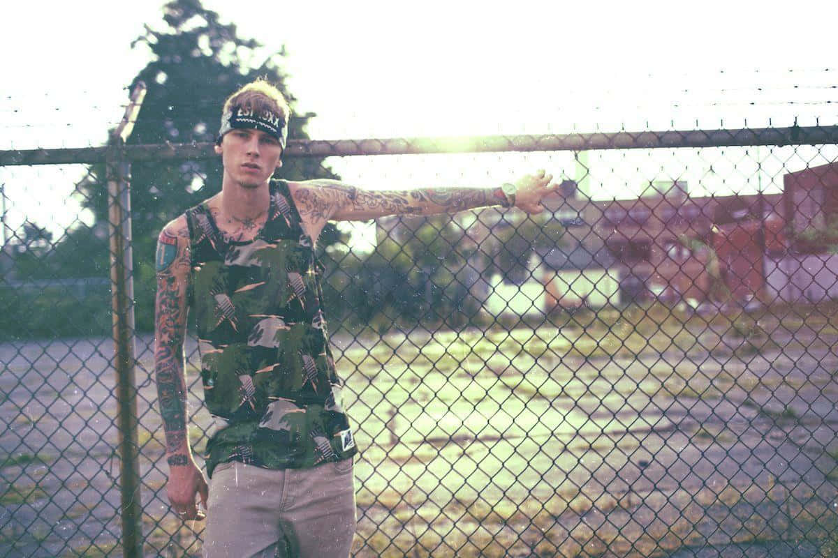 mgk 桌布 壁紙 — 一位紋身年輕男子穿著迷彩背心，靠著鐵絲網圍欄，身後有太陽眩光。