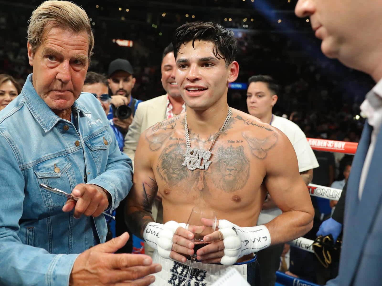 Ryan Garcia 桌布 壁紙 — 一位紋身拳擊手戴著白色手套，在燈光明亮的拳擊場內微笑著舉起酒杯，周圍都是人。