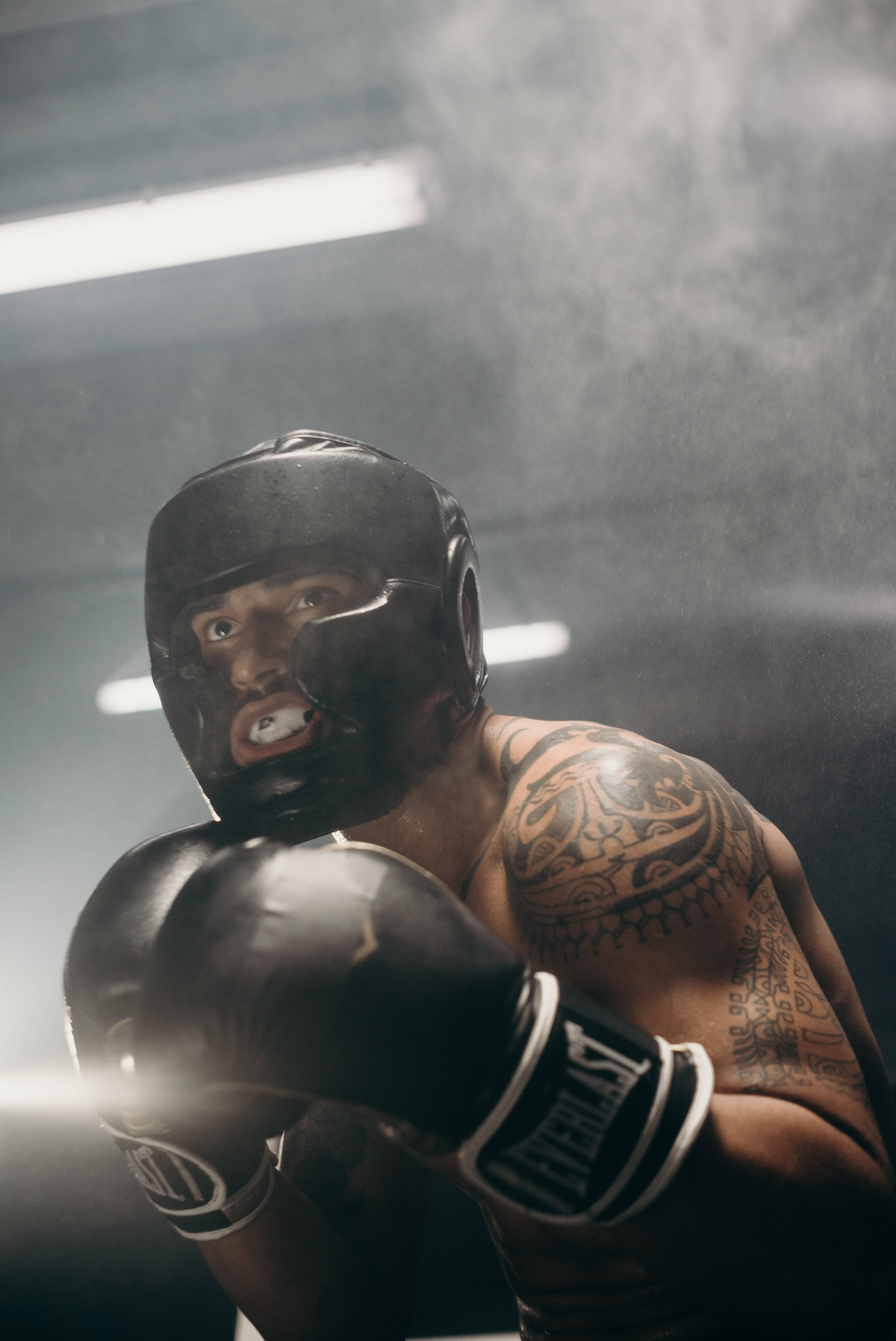 Cool Boxing 桌布 壁紙 — 一位紋身拳擊手穿著黑色裝備，在明亮的健身房燈光下透過煙霧凝視。