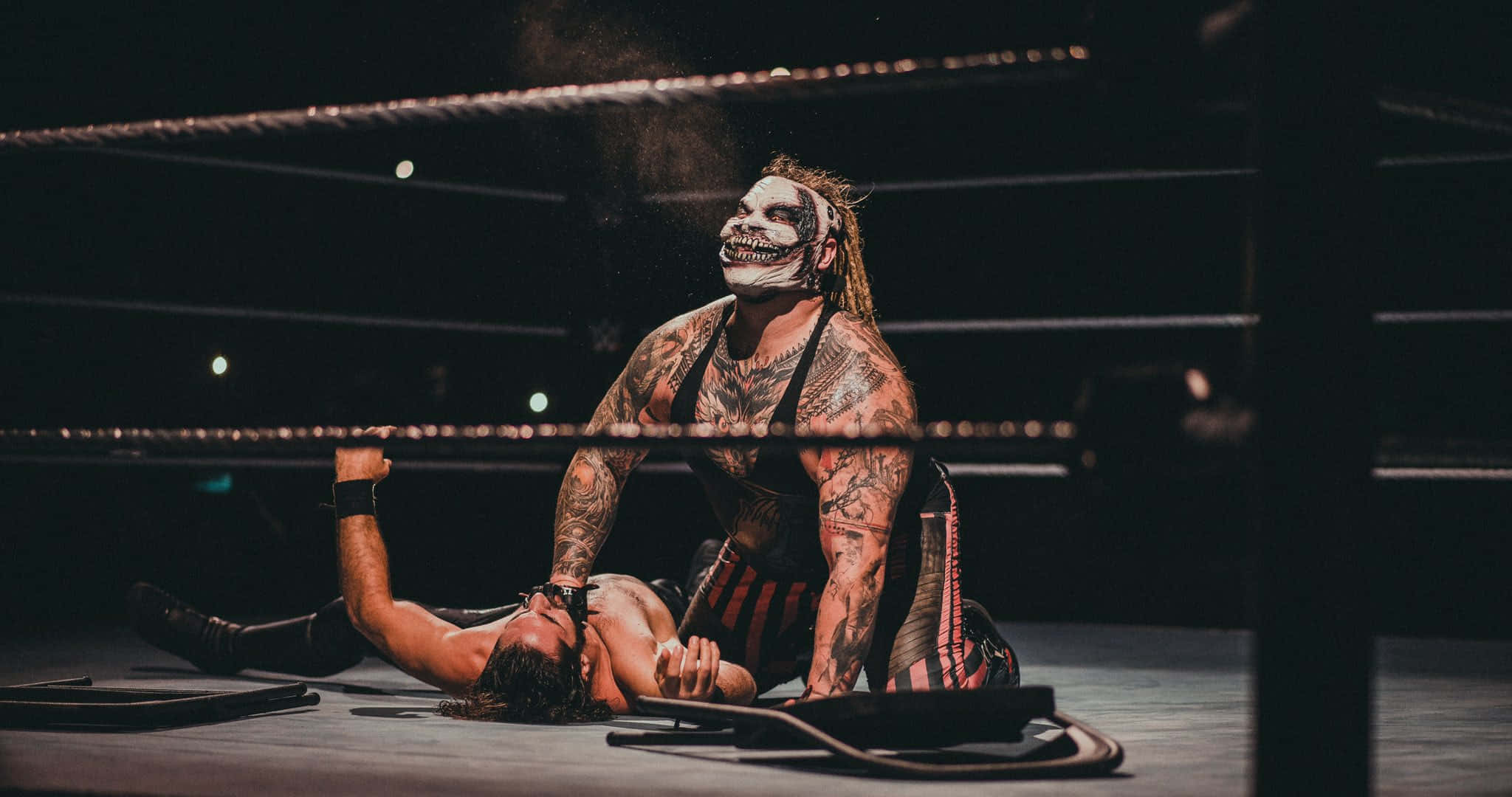 WWE 桌布 壁紙 — 一位紋身摔角手戴著骷髏面具，在昏暗的摔角擂台上將對手壓制在墊子上。
