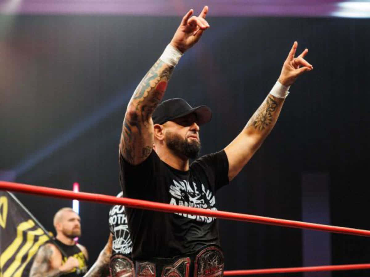 WWE 桌布 壁紙 — 一位紋身摔角手身穿黑色襯衫和帽子，在紅色擂台上高舉雙手慶祝勝利，並持有冠軍腰帶。