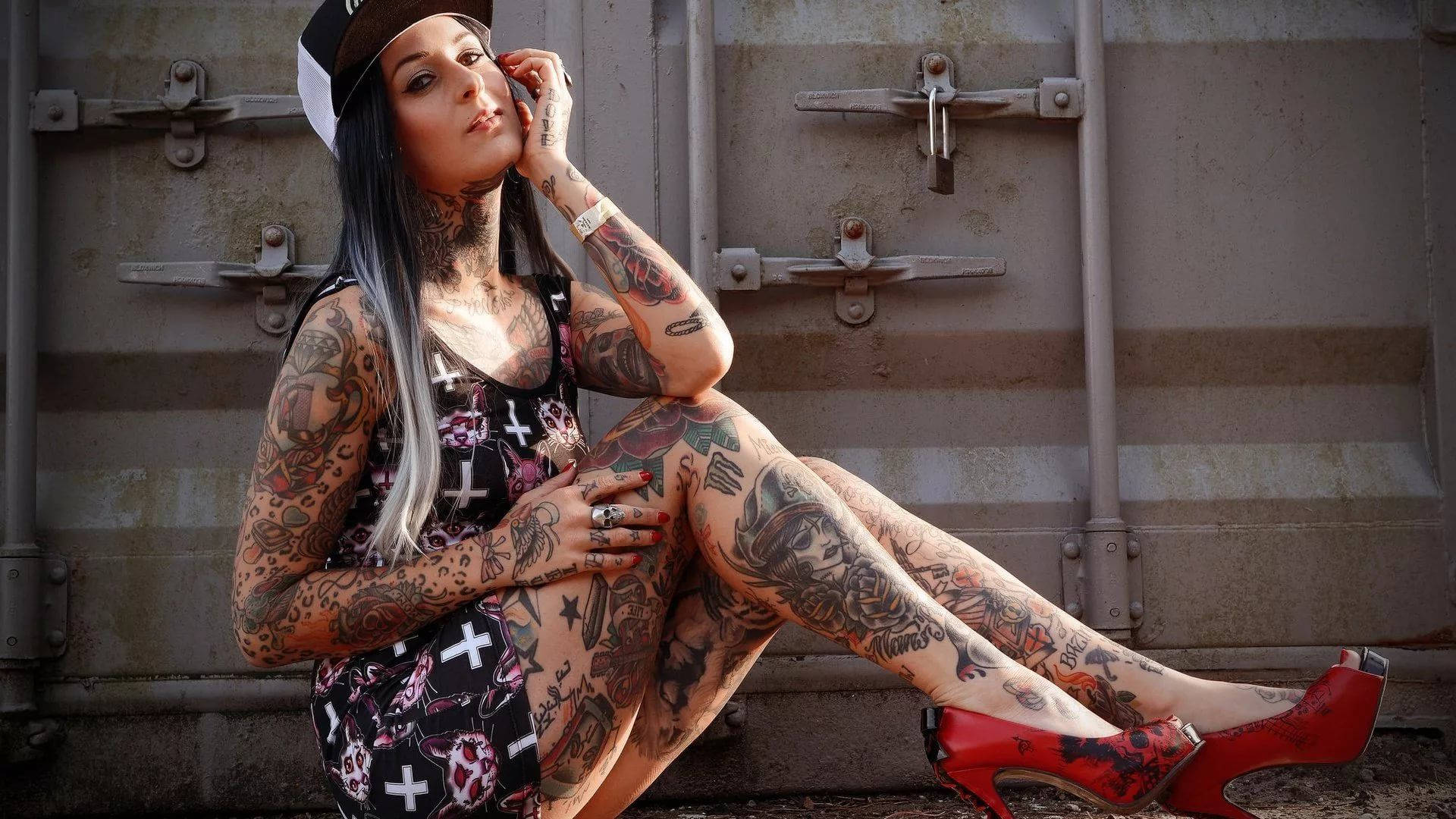 Tattoo 桌布 壁紙 — 一位紋身濃密的女性穿著紅色高跟鞋，在風化的金屬貨櫃前自信地擺姿勢。