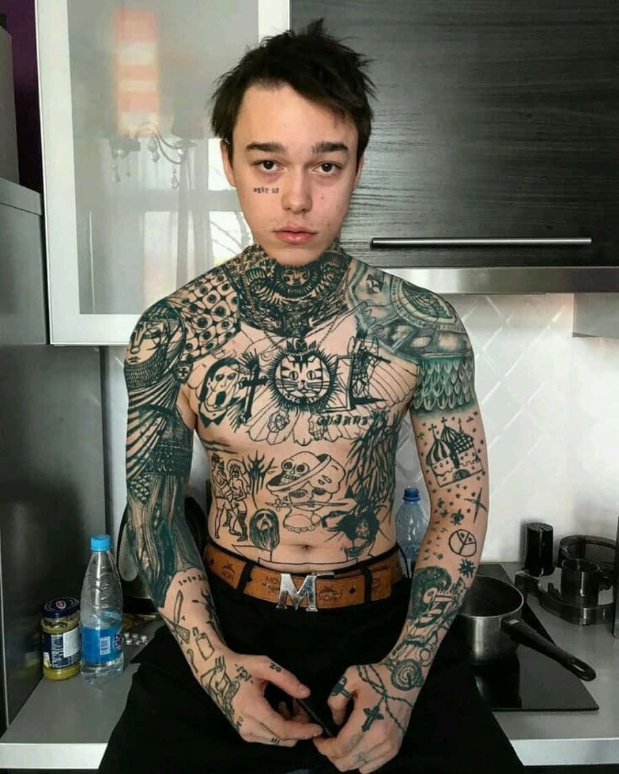 Tattoo Boy 桌布 壁紙 — 一位紋身濃密的年輕男子，深色頭髮，表情嚴肅，站在現代廚房裡，手臂和軀幹佈滿複雜的黑色墨水圖案。