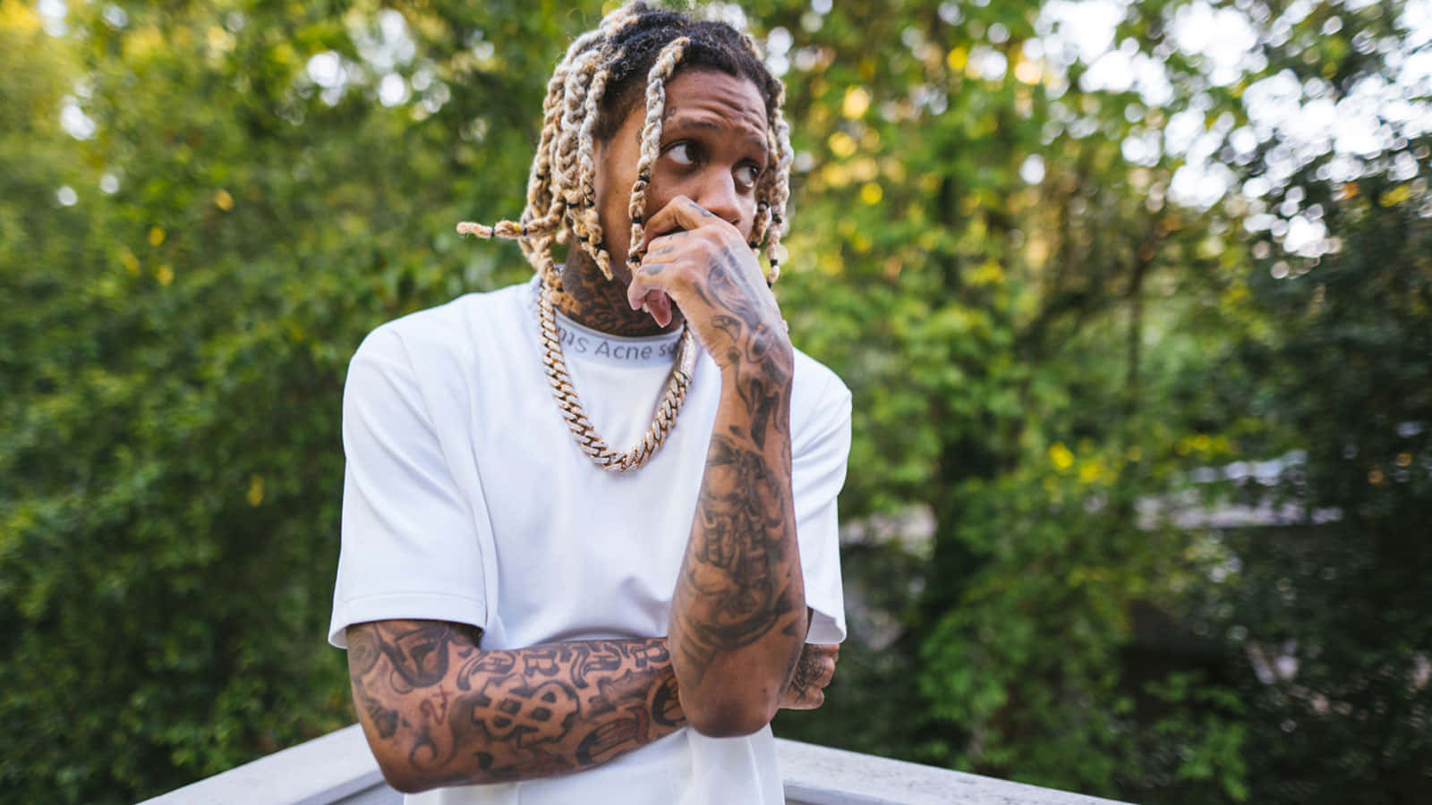 Lil Durk 桌布 壁紙 — 一位紋身男子，辮子頭，身穿白色T恤，靠在欄杆上，在茂密的綠葉中若有所思地凝視。
