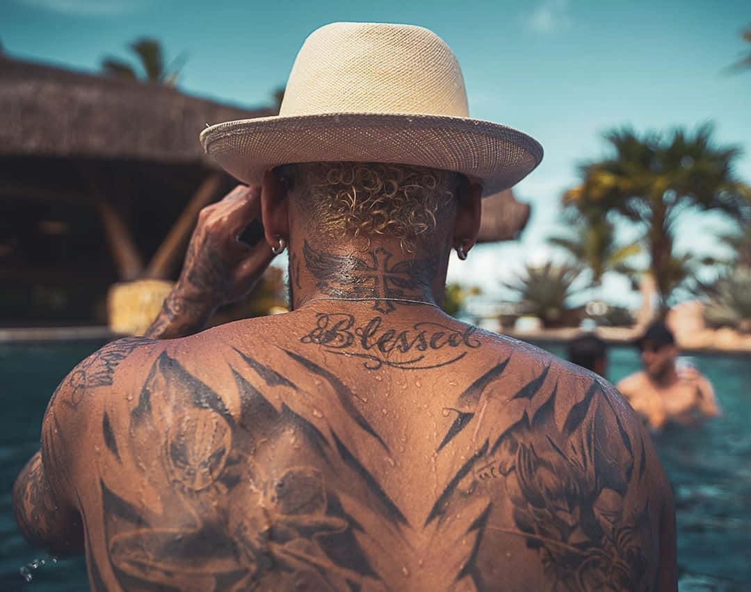 Tattoo 桌布 壁紙 — 一位紋身男子戴著草帽，站在泳池旁，背景有棕櫚樹和茅草屋頂。