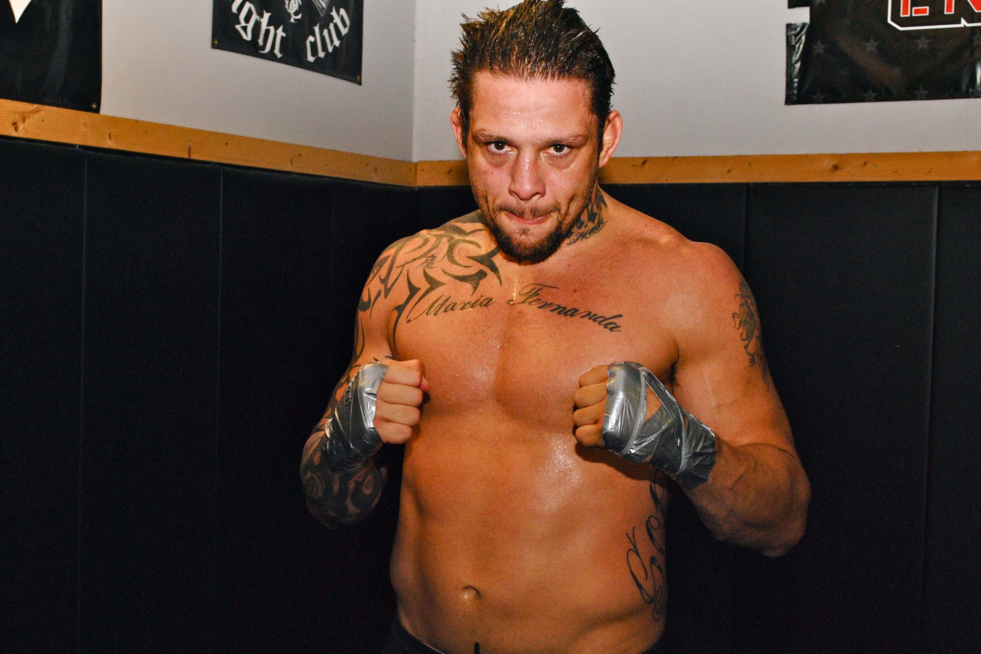 MMA 桌布 壁紙 — 一位紋身男性拳擊手戴著銀色護手繃帶，在健身房角落凝視鏡頭。
