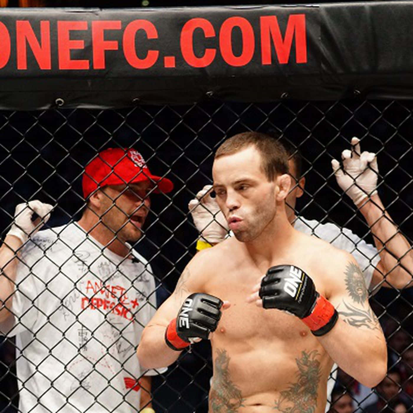 martial arts 桌布 壁紙 — 一位紋身的MMA拳手戴著黑色和紅色手套站在八角籠內，身後有一位戴紅色帽子的教練在為他打氣。