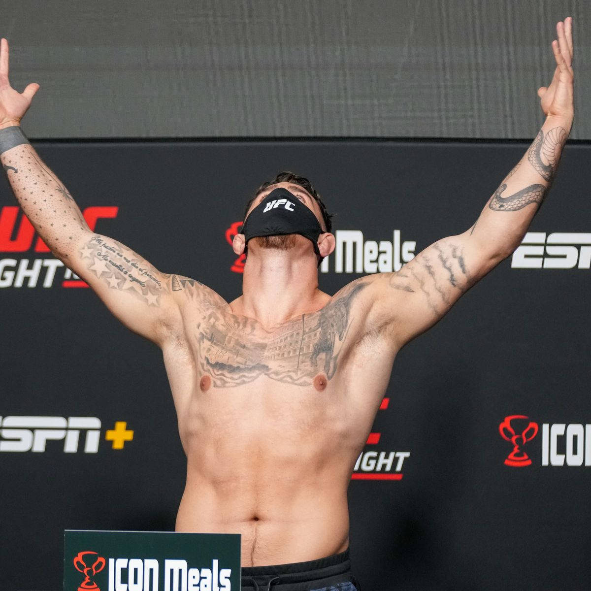 UFC 桌布 壁紙 — 一位紋身的UFC拳手在稱重活動中勝利地高舉雙臂，戴著黑色品牌口罩。