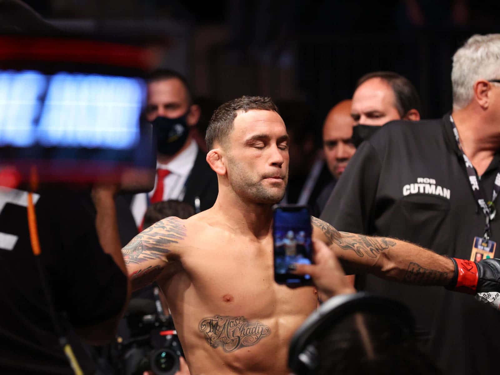 UFC 桌布 壁紙 — 一位紋身的戰士在昏暗的競技場赤裸上身站著，粉絲和工作人員用手機和相機包圍他。