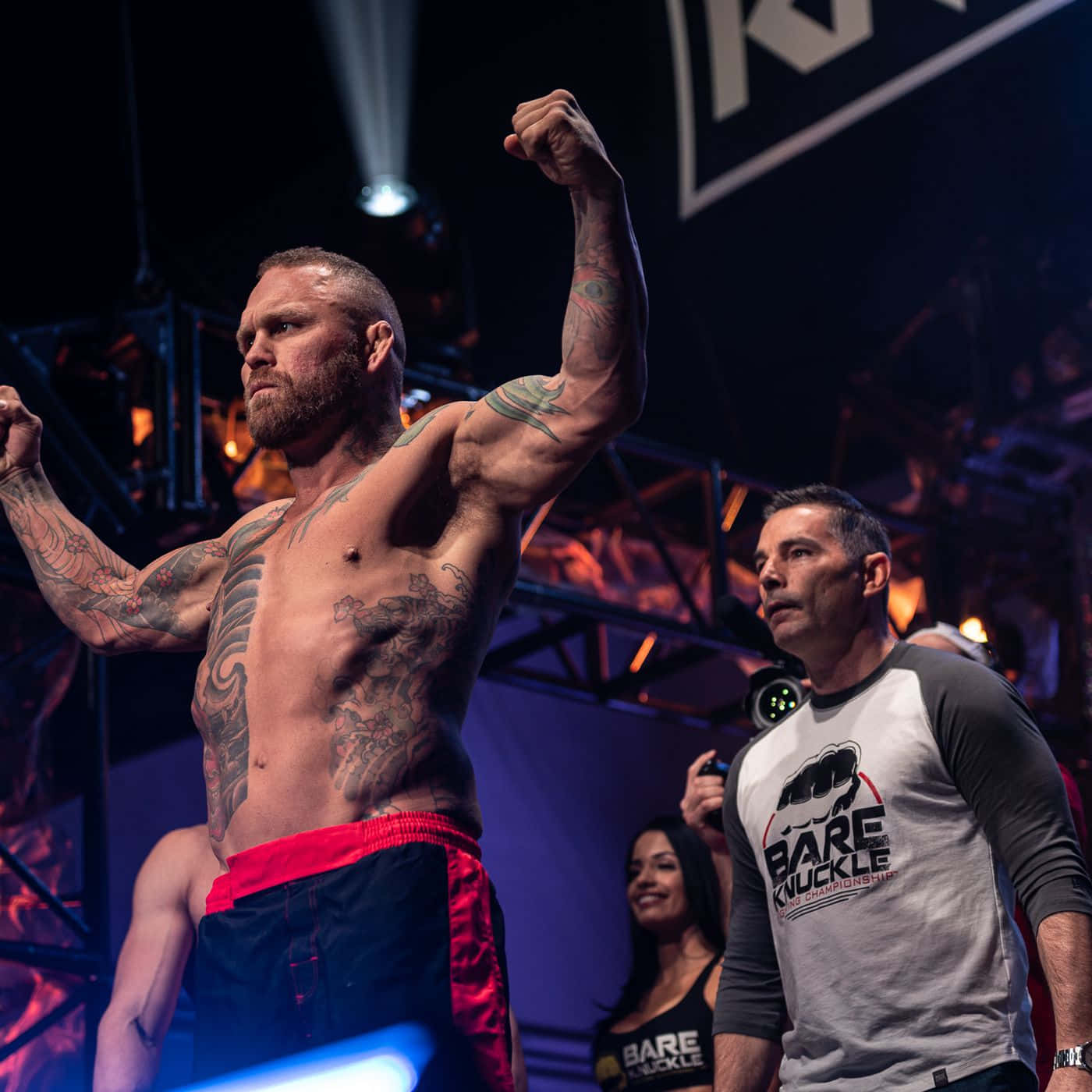 MMA 桌布 壁紙 — 一位紋身的裸拳拳擊手在競技場燈光下，背景有裁判和粉絲，勝利地舉起拳頭。