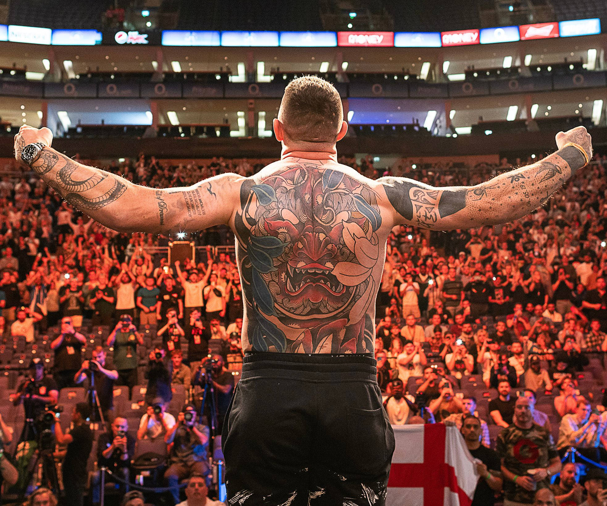 UFC 桌布 壁紙 — 一位紋身運動員在燈光明亮的競技場上，高舉雙臂，在歡呼的人群面前慶祝勝利。