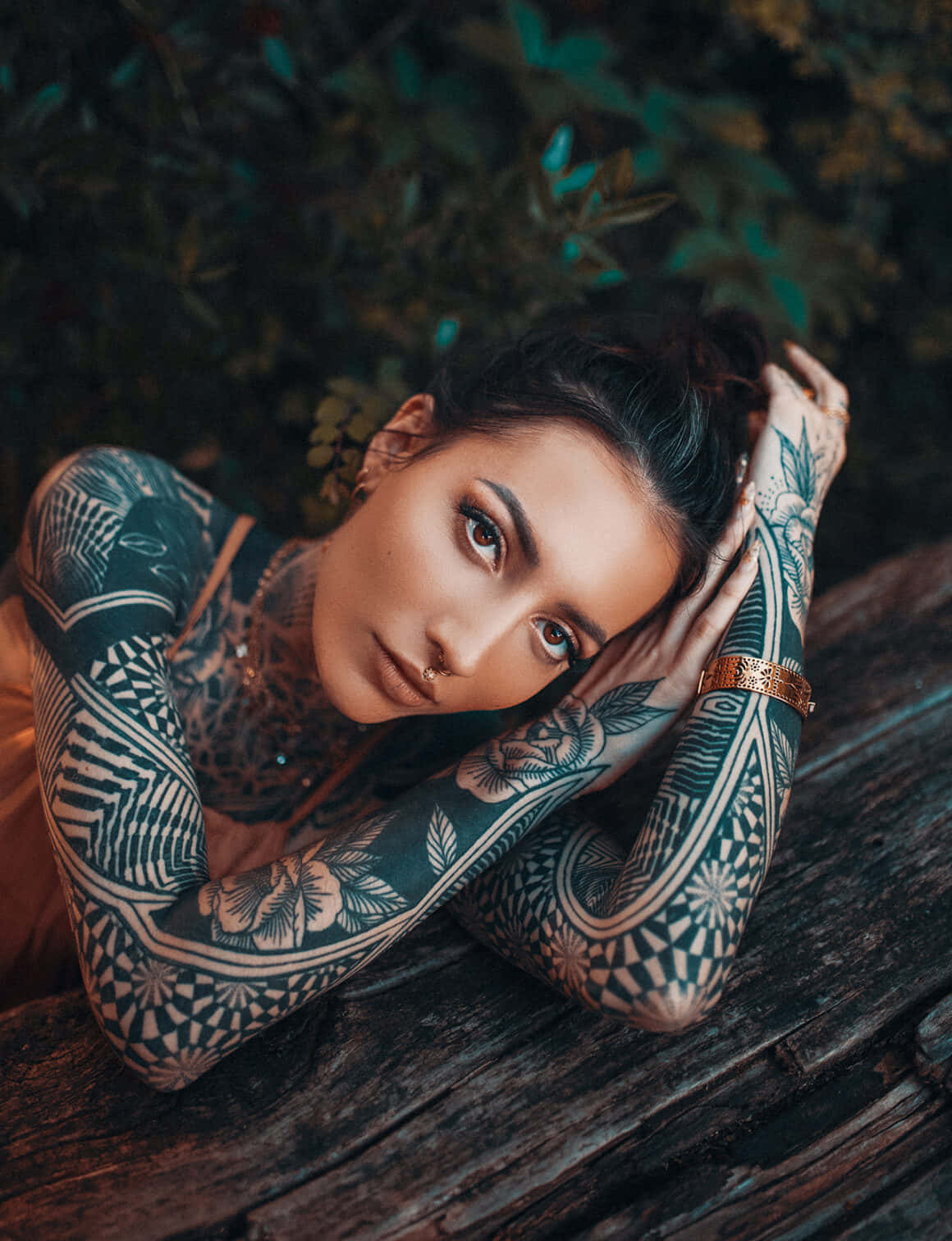 Tattoo Girl 桌布 壁紙 — 一位紋身黑髮女子將頭靠在手臂上，置於風化木頭上，周圍環繞著茂密的綠色樹葉。
