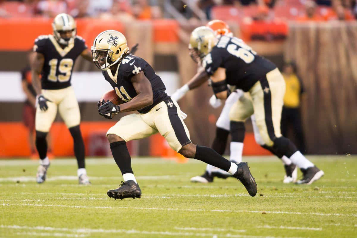 Alvin Kamara 桌布 壁紙 — 一位紐奧良聖徒隊的美式足球員身穿黑金色球衣，持球衝刺，隊友和對手在充滿活力的綠色球場上追趕。