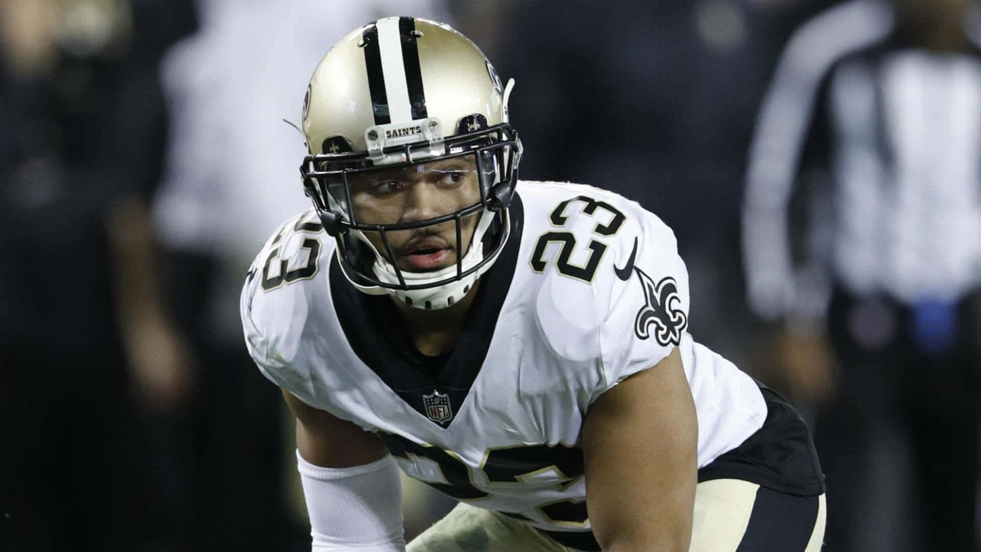 New Orleans Saints 桌布 壁紙 — 一位紐奧良聖徒隊的美式足球員身穿白色和金色球衣，在球場燈光下的夜間比賽中激烈地蹲伏。