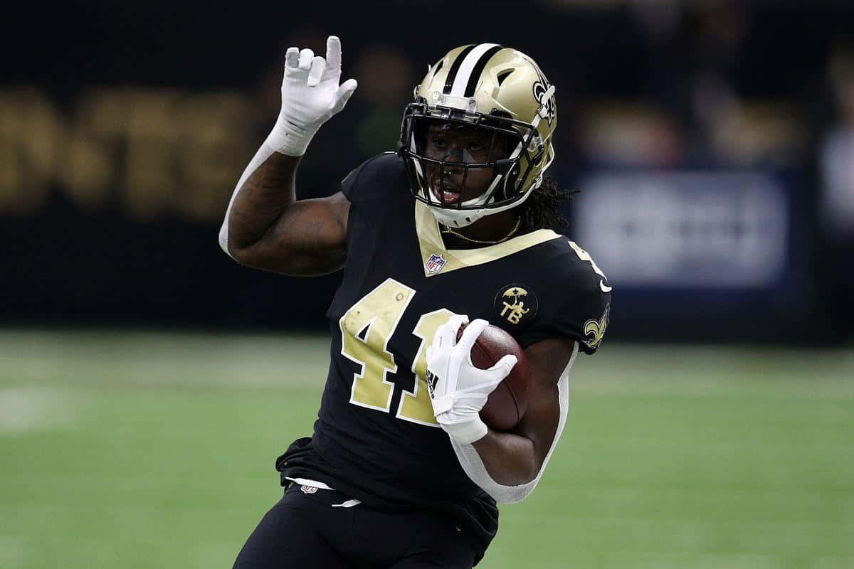 Alvin Kamara 桌布 壁紙 — 一位紐奧良聖徒隊的美式足球員身穿黑金色球衣，帶著球奔跑並舉手慶祝，在綠色球場上。