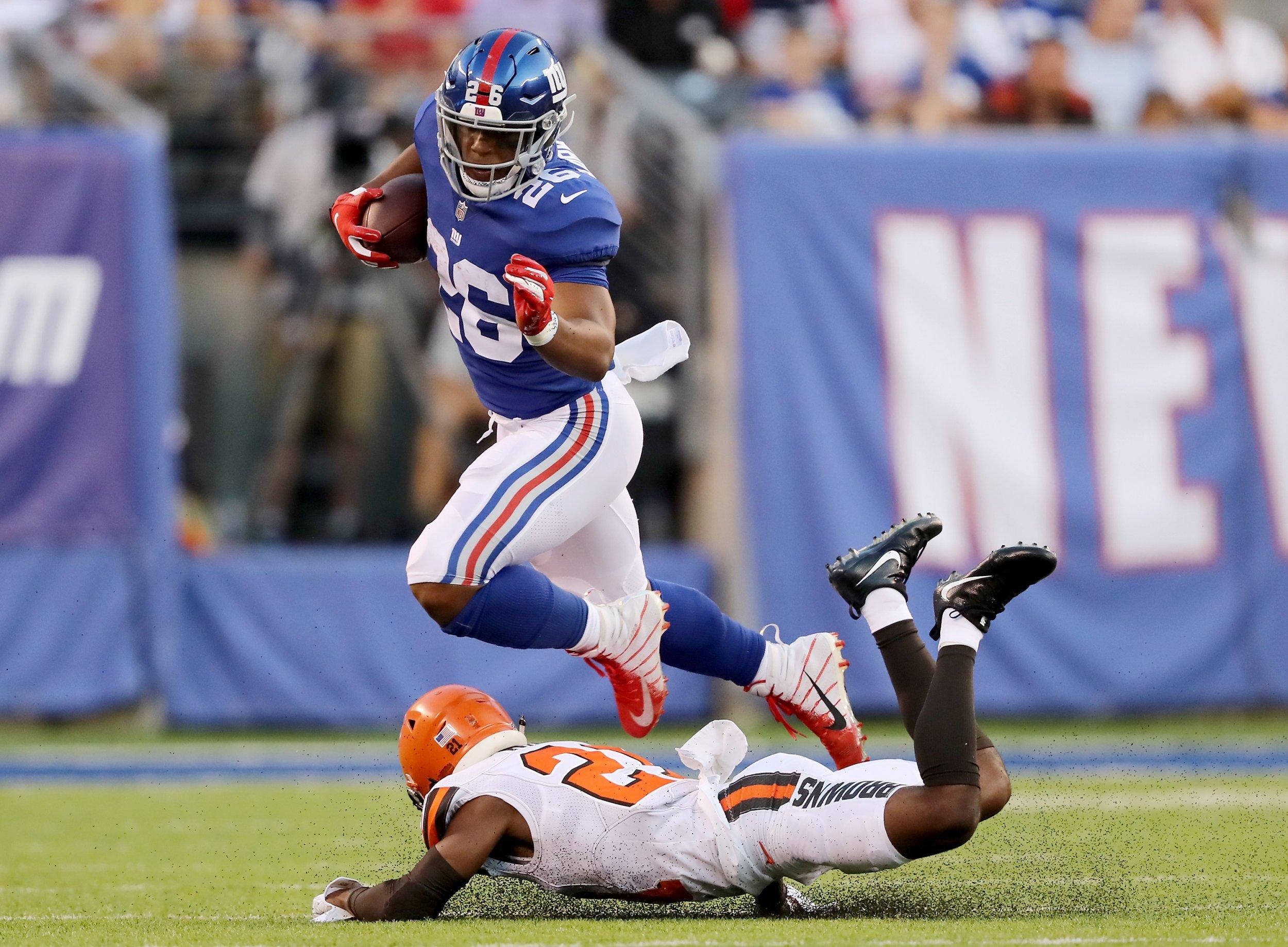 Saquon Barkley 桌布 壁紙 — 一位紐約巨人隊球員在日間比賽中，跳過一名倒地的克里夫蘭布朗隊防守球員，同時緊緊抓住橄欖球。