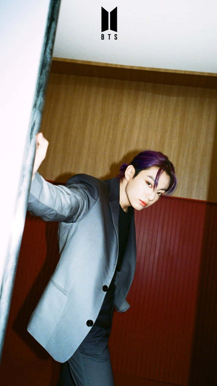 Jungkook 桌布 壁紙 — 一位紫髮年輕男子穿著灰色西裝，靠著溫暖的木質和紅色鑲板牆。