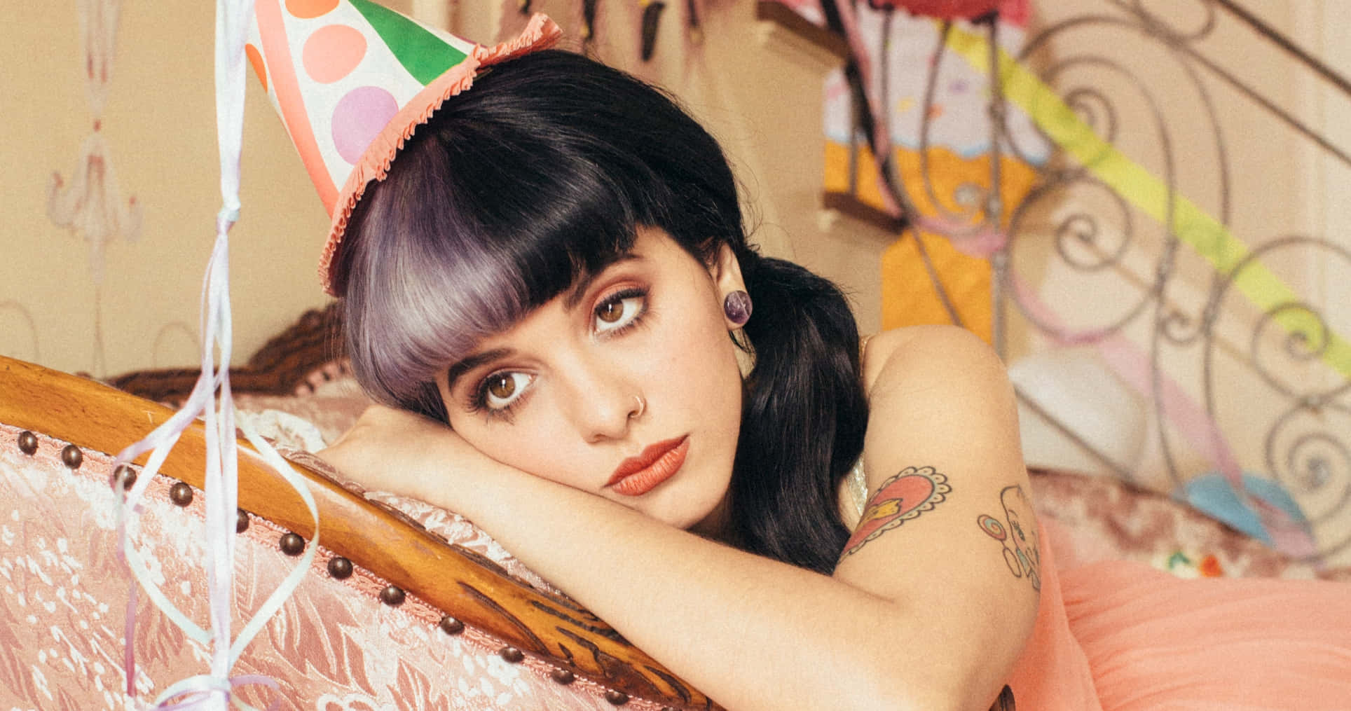 Melanie Martinez Aesthetic 桌布 壁紙 — 一位紫髮戴派對帽的年輕女子，將頭靠在帶有彩色漩渦和珠子的復古床上。