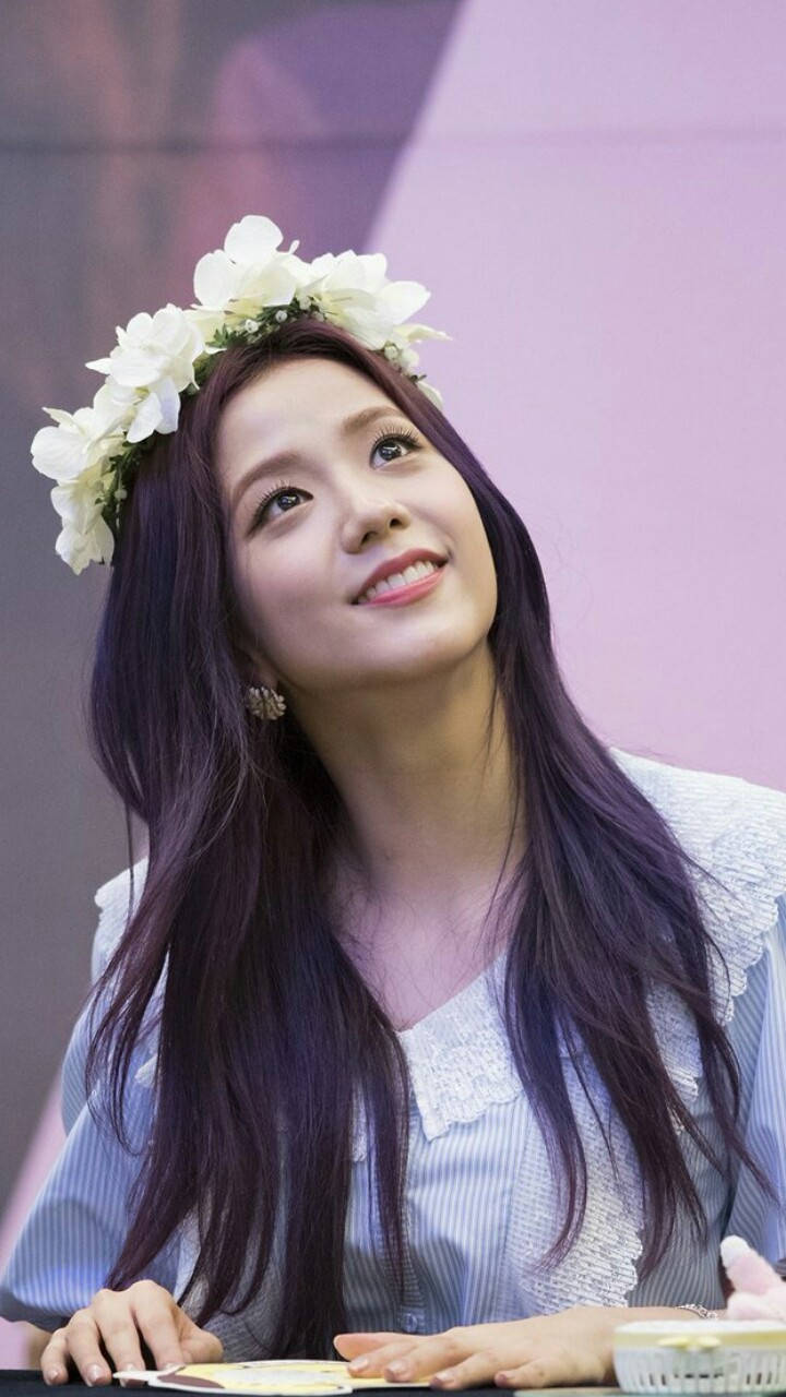 Jisoo Cute 桌布 壁紙 — 一位紫髮戴白色花冠的年輕女子坐在桌旁溫柔微笑。