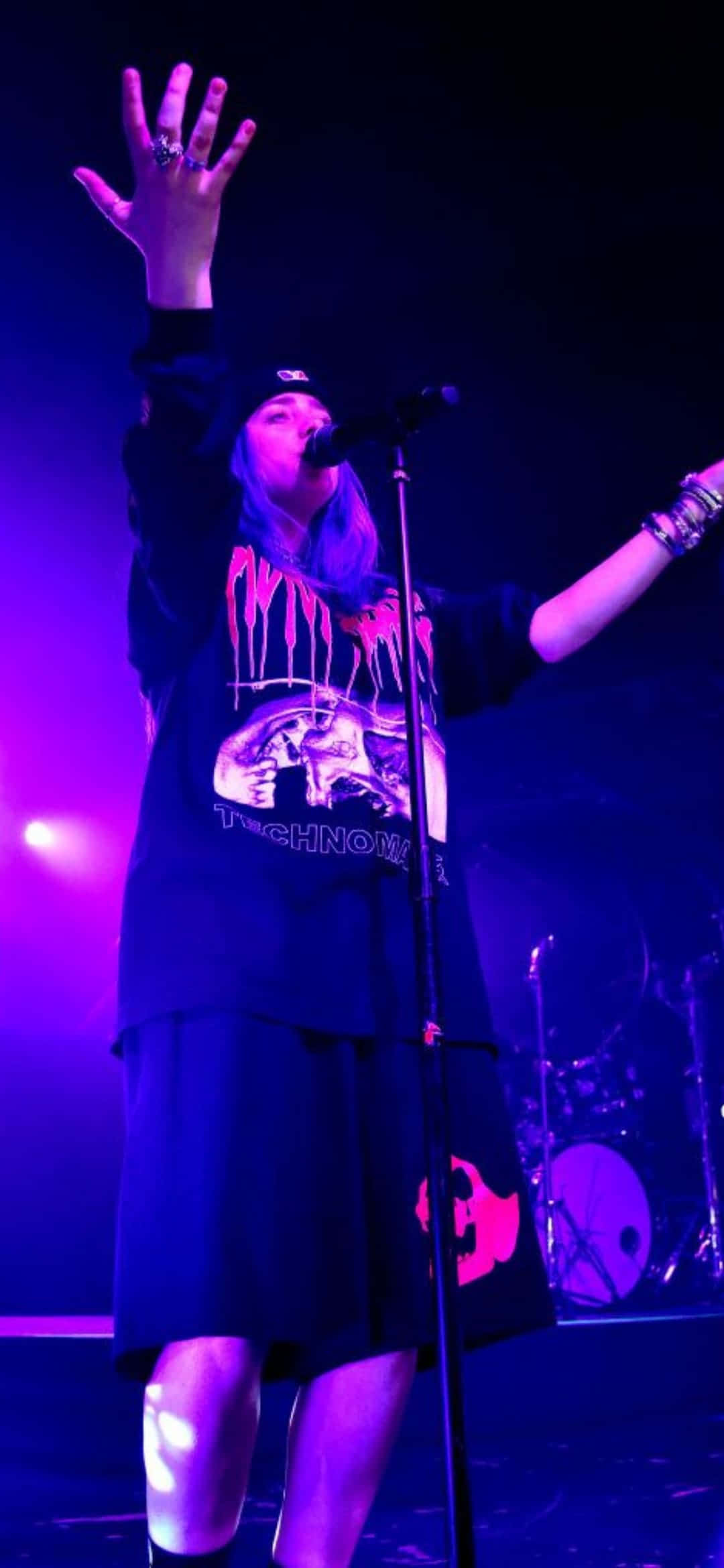 Billie Eilish Purple 桌布 壁紙 — 一位紫髮表演者在充滿活力的紫色燈光下高舉雙臂熱情歌唱。