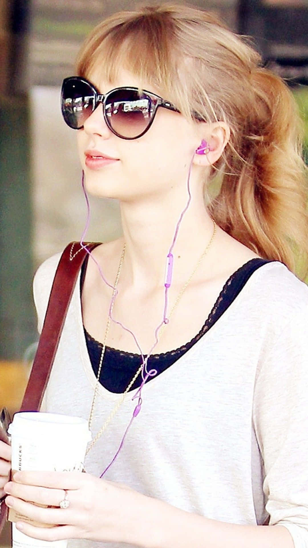 Taylor Swift iPhone 桌布 壁紙 — 一位紮著馬尾辮的金髮年輕女子戴著太陽眼鏡和紫色耳機，手持咖啡杯。