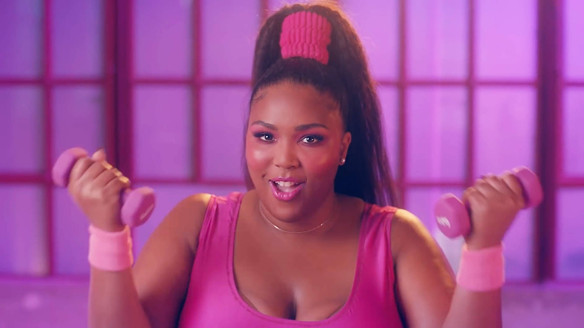 Lizzo 桌布 壁紙 — 一位紮著高馬尾、佩戴粉色配飾的女性，在紫色鑲板背景前微笑著舉起粉色啞鈴。