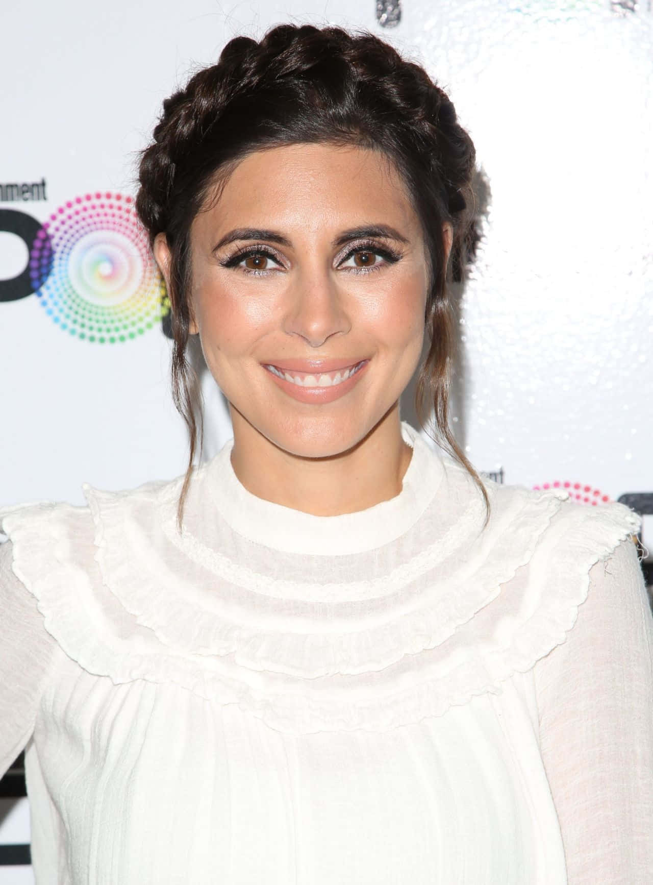 Jamie Lynn Sigler 桌布 壁紙 — 一位紮辮子的微笑女士，穿著白色荷葉邊襯衫，背景是色彩繽紛的活動場景。