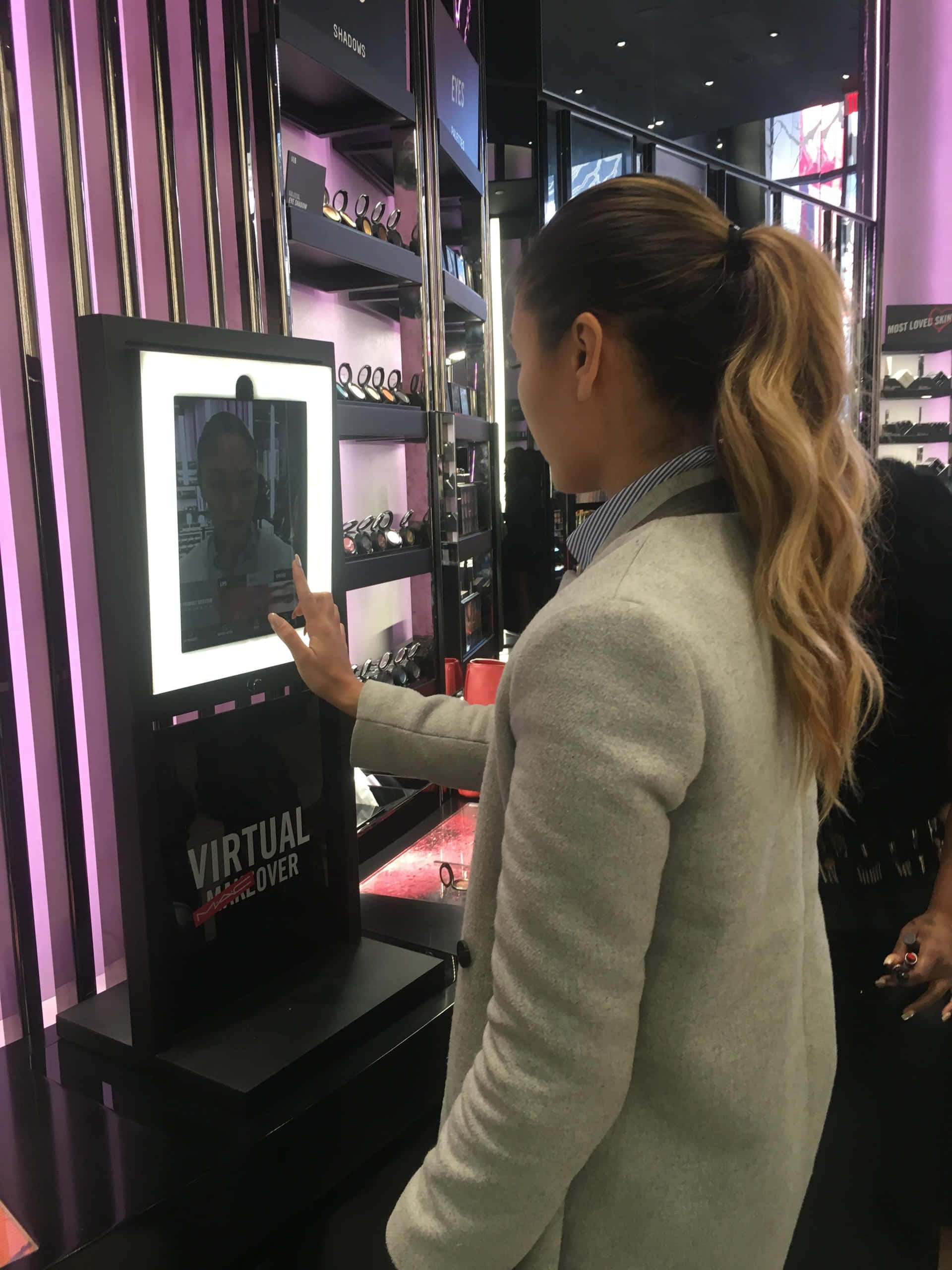 mirror 桌布 壁紙 — 一位紮馬尾的女士正在現代零售店內與虛擬化妝鏡互動，店內有紫色燈光和產品貨架。