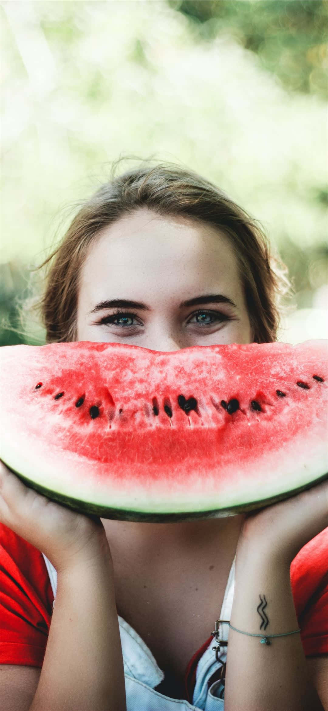 Watermelon iPhone 桌布 壁紙 — 一位綠眼睛的年輕女子頑皮地從她舉在臉前的一大塊多汁西瓜片上方偷看。