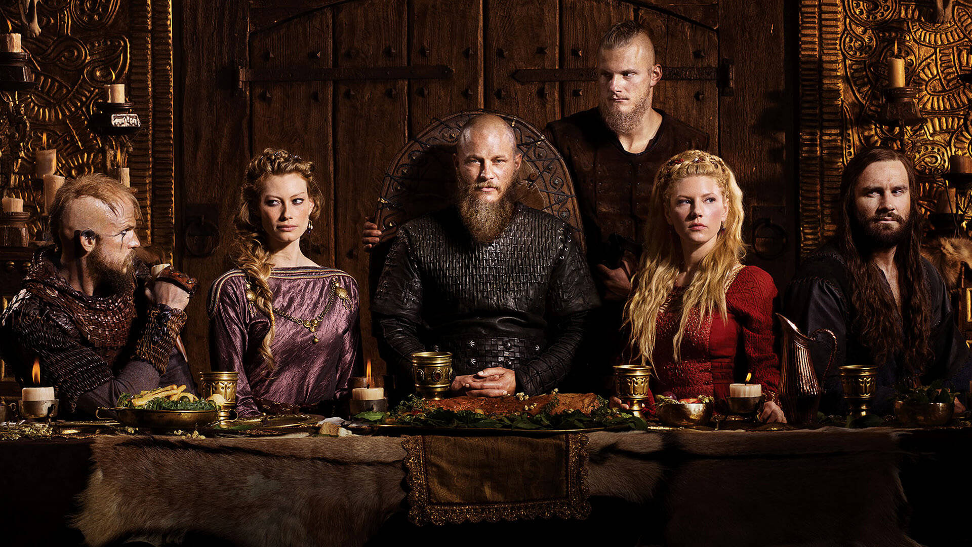Vikings 桌布 壁紙 — 一位維京首領和他的船員莊嚴地坐在一個裝飾著複雜雕刻的黑暗木製大廳裡，享用燭光盛宴。