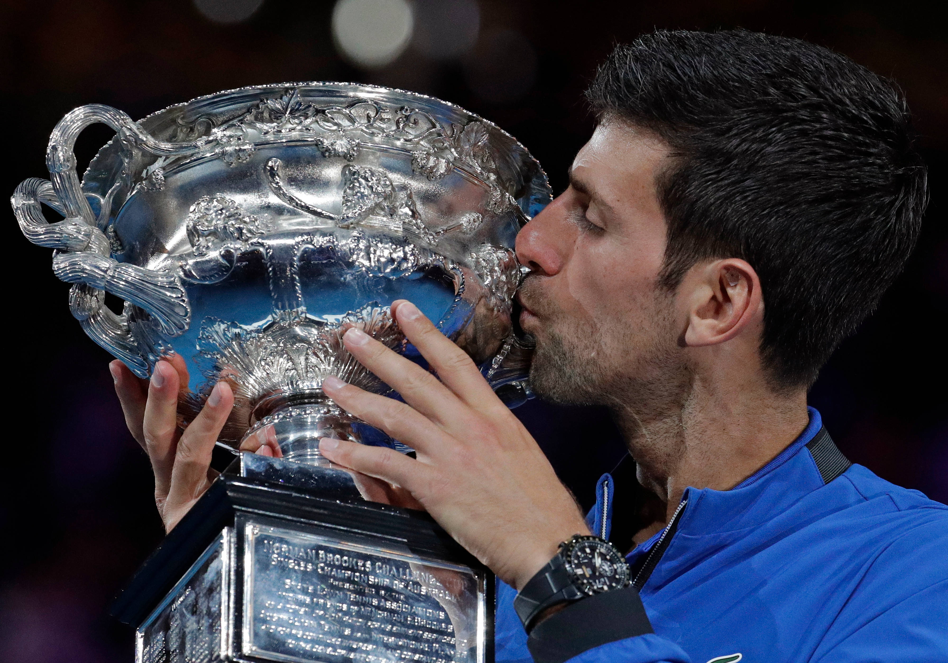 Novak 桌布 壁紙 — 一位網球運動員在明亮的體育場燈光下親吻一個帶有複雜獅子圖案的大型華麗銀色獎盃。
