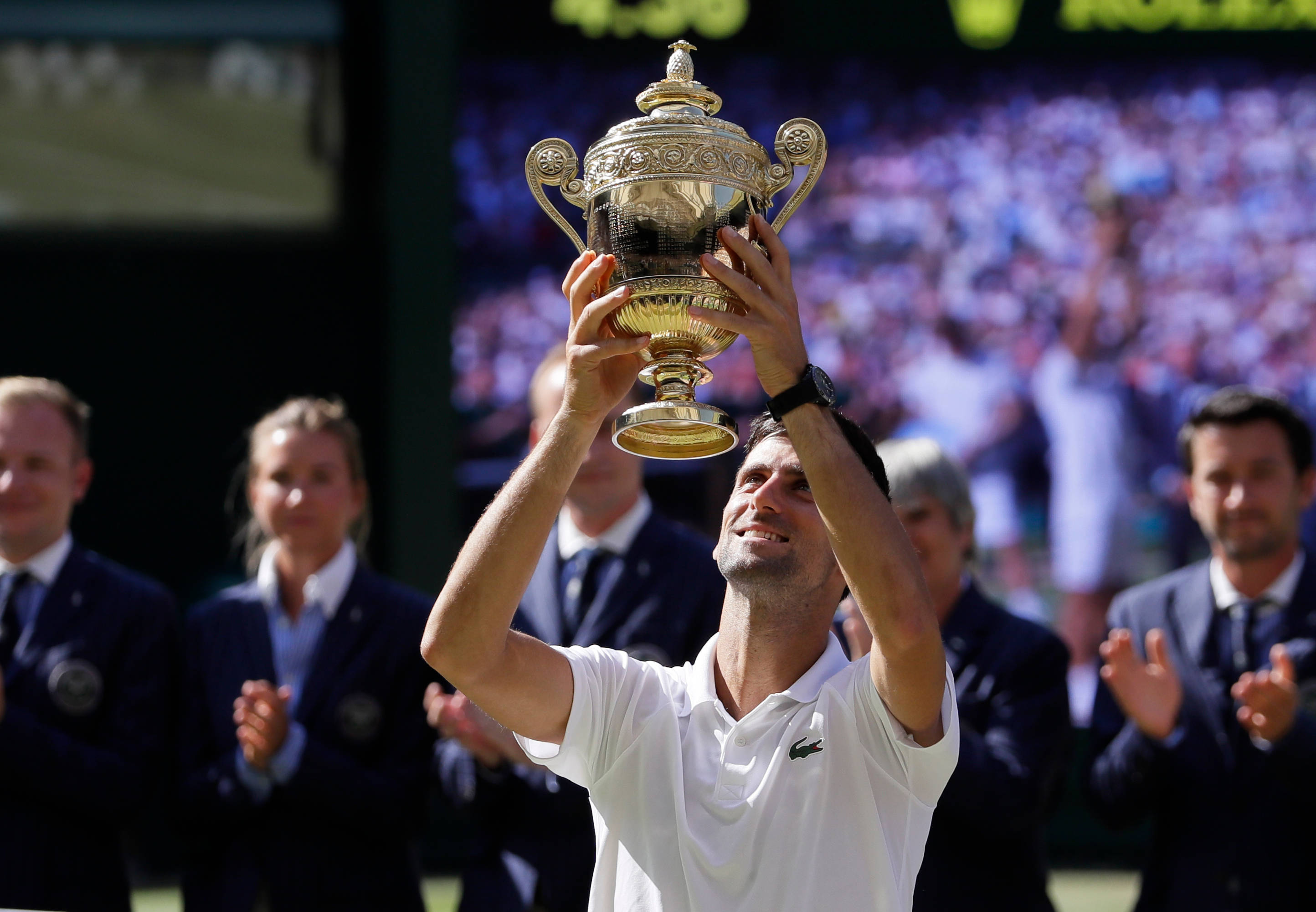 Novak 桌布 壁紙 — 一位網球運動員在陽光明媚的草地上，在觀眾的掌聲中，勝利地將閃閃發光的金色獎盃高舉過頭。