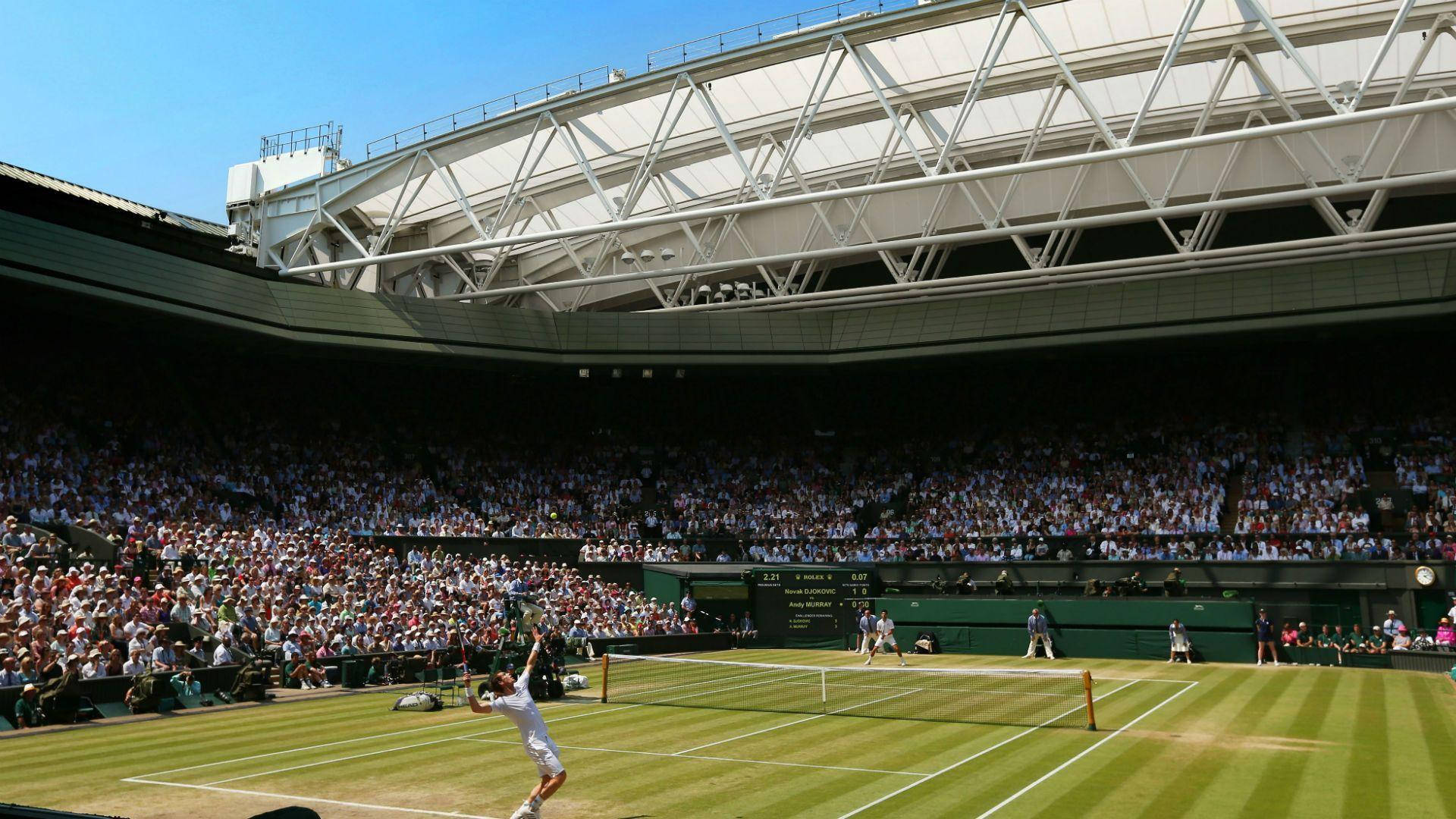 Wimbledon 桌布 壁紙 — 一位網球選手在明亮的藍天下，於草地場上跳起發球，滿座的體育場正在觀看。