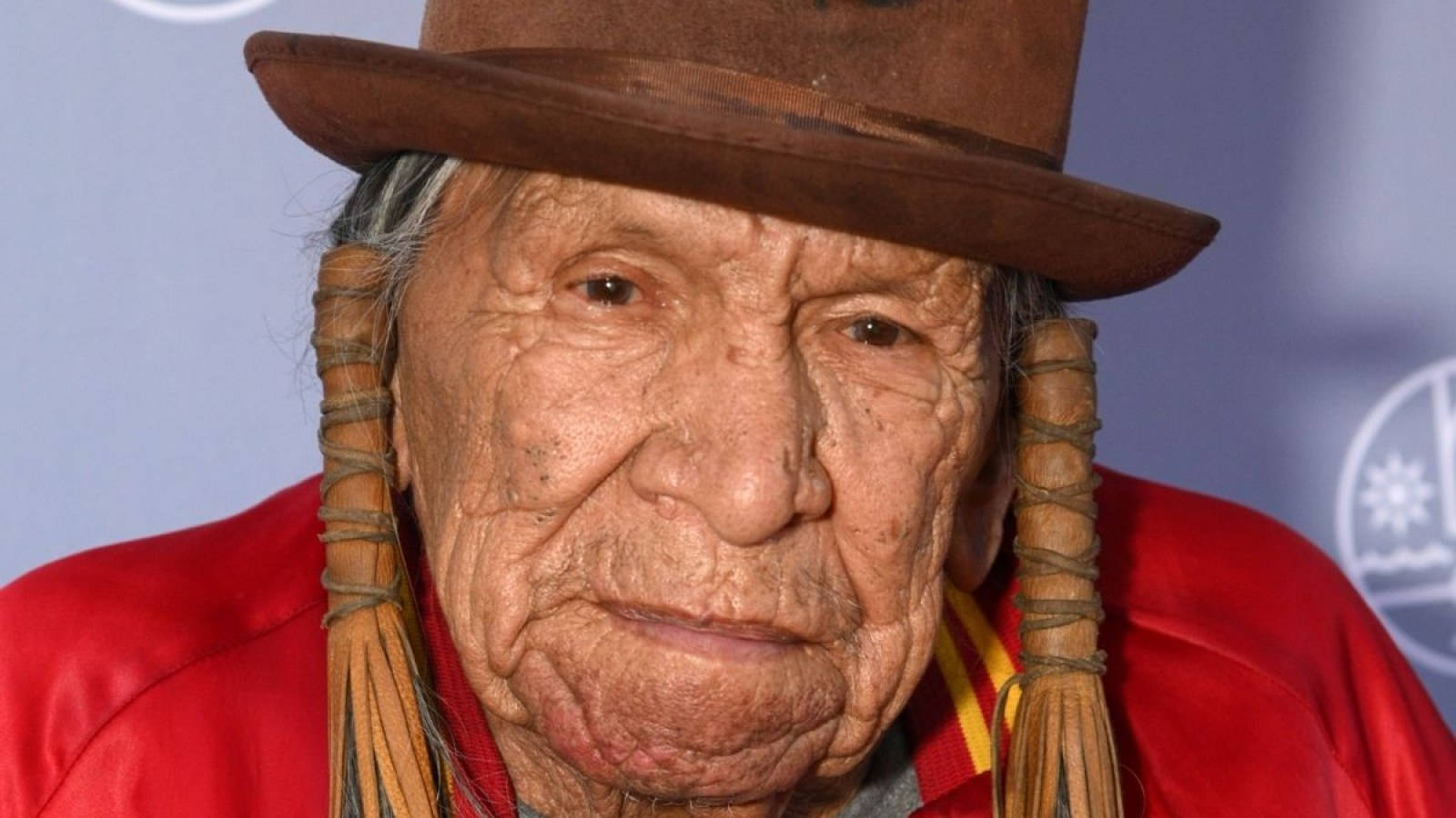 Native American 桌布 壁紙 — 一位編辮子的老年美洲原住民男子戴棕色帽子和紅色外套的特寫肖像。