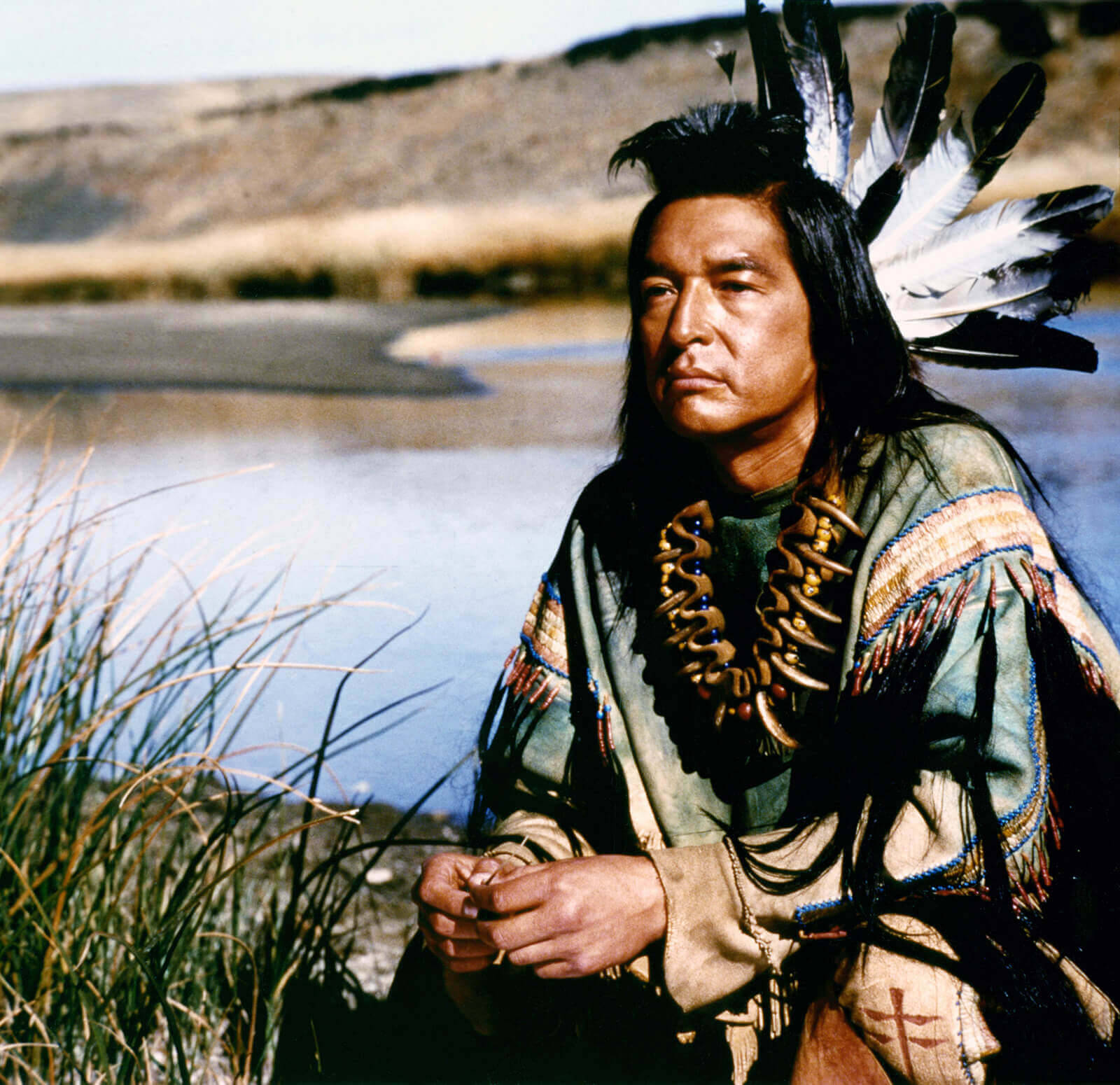 Native American 桌布 壁紙 — 一位美洲原住民男子，身穿傳統羽毛頭飾和珠飾服裝，沉思地坐在平靜的河岸邊。