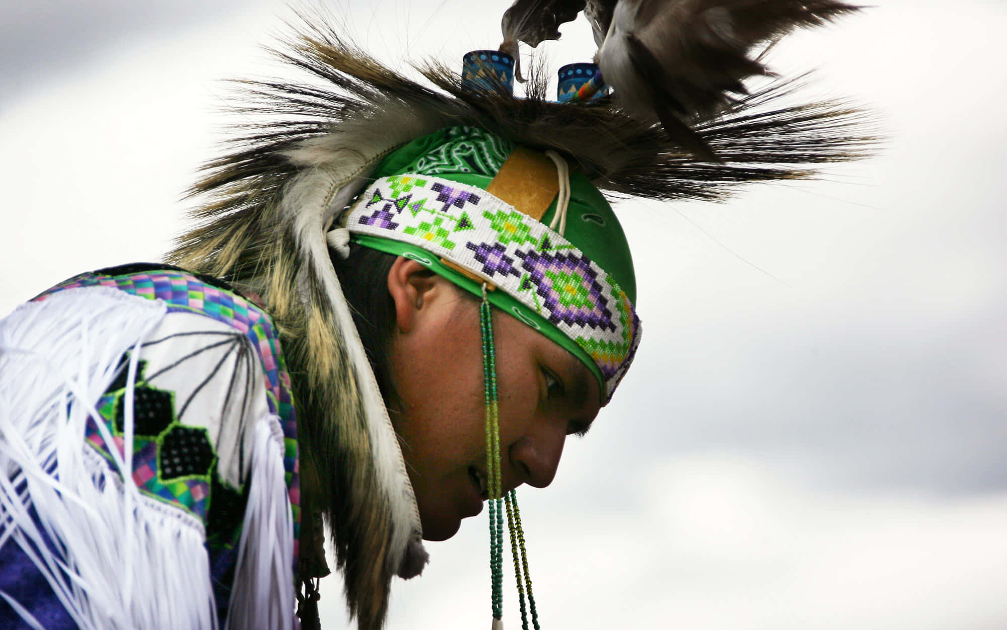 Native American 桌布 壁紙 — 一位美洲原住民男子，頭戴傳統羽毛頭飾，身穿珠飾禮服，在陰雲密布的天空下低頭凝視。