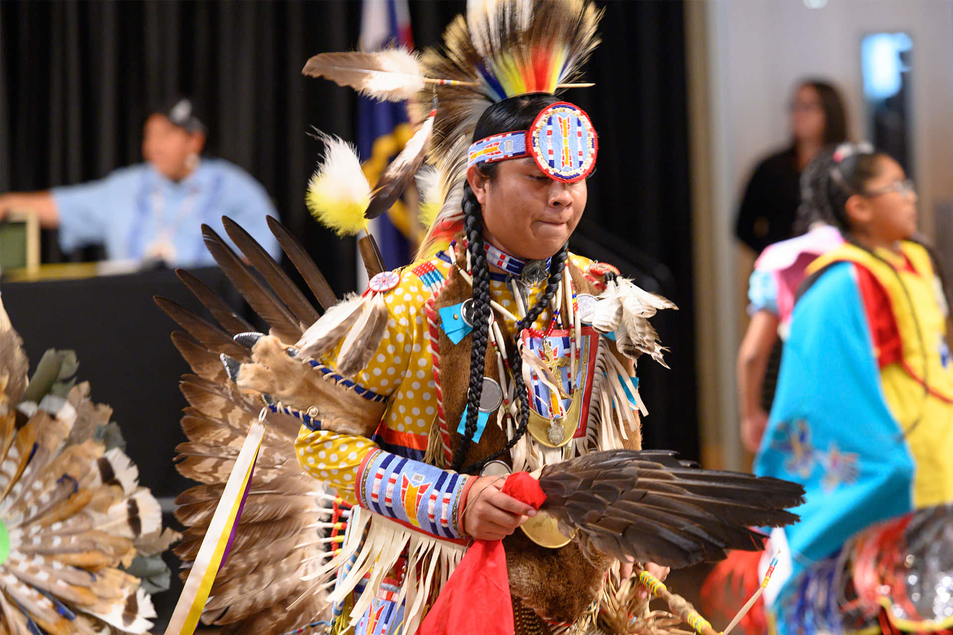Native American 桌布 壁紙 — 一位美洲原住民舞者身著鮮豔的禮服，配戴鷹羽和珠飾頭飾，在室內文化儀式中表演。