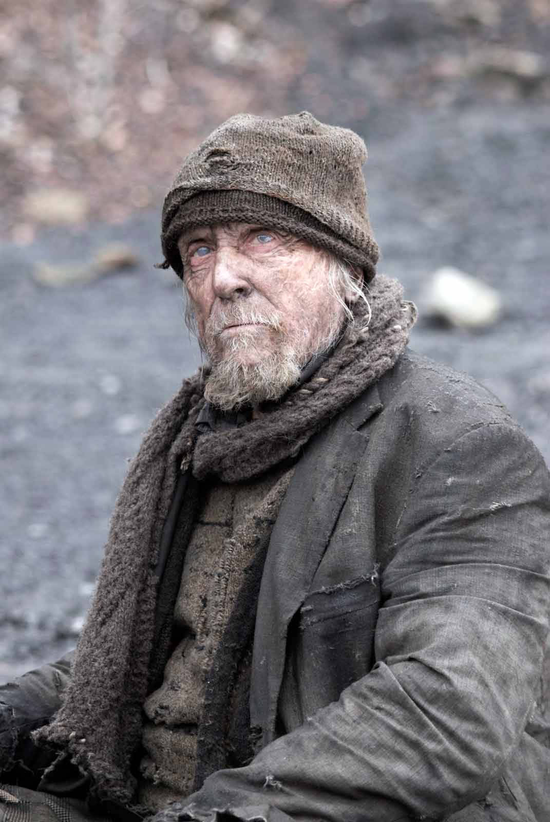 Celebrity 桌布 壁紙 — 一位老人，擁有銳利的藍眼睛和飽經風霜的臉龐，坐在荒涼的岩石景觀中，穿著破舊的羊毛衣服。