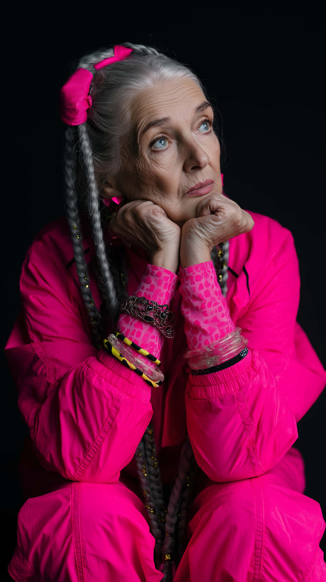 Expressive Senior Woman in Vibrant Modern Fashion Portrait 壁紙 — 一位老婦人，灰色辮子繫著粉紅色蝴蝶結，托腮仰望，背景為黑色。