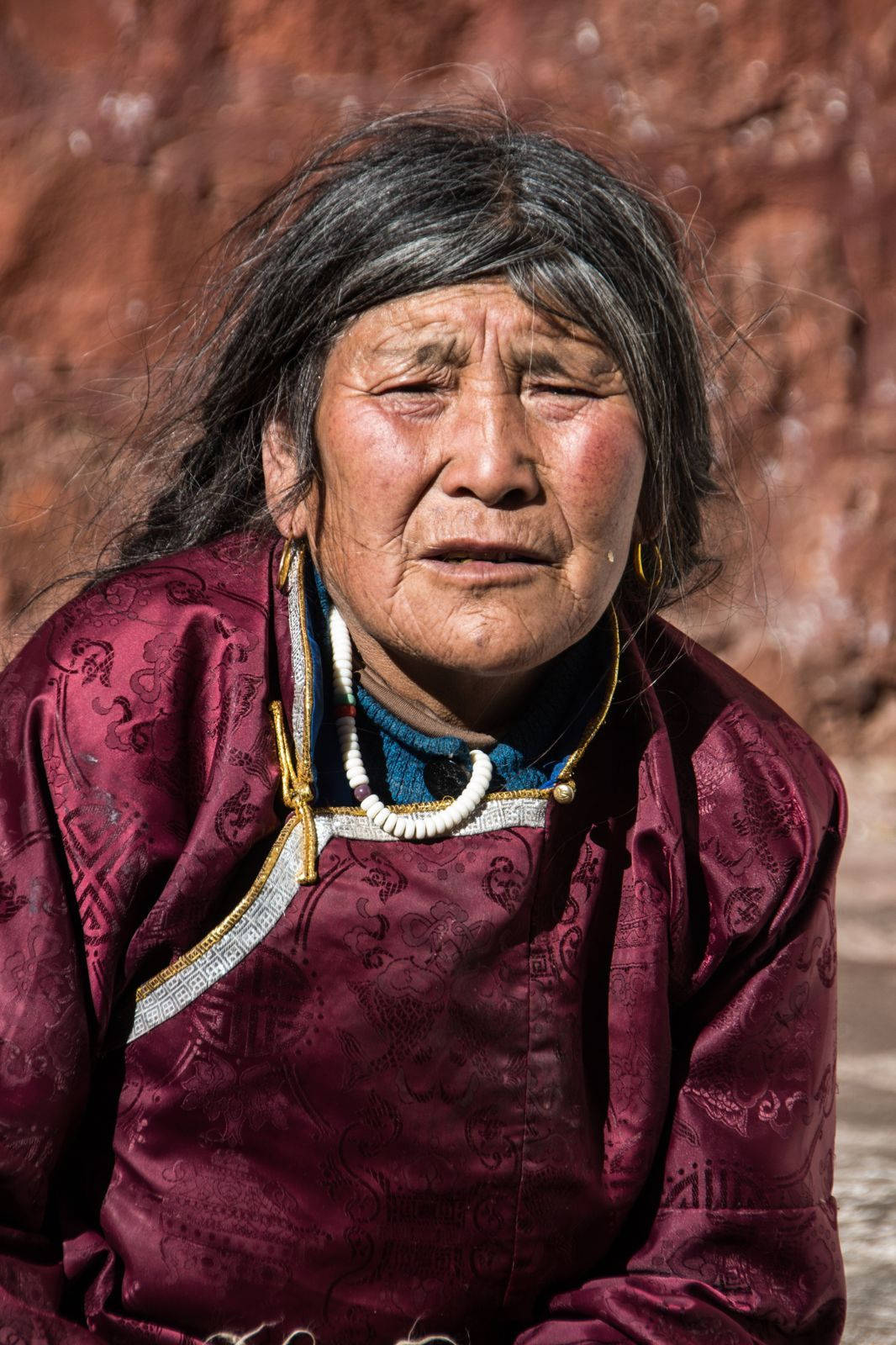 Woman 桌布 壁紙 — 一位老婦人，面容飽經風霜，深色頭髮，身穿栗色圖案長袍，背景是紅色岩石。