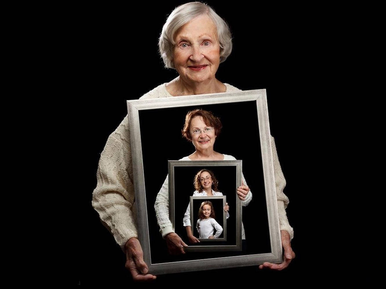 Grandma 桌布 壁紙 — 一位老婦人微笑著，手持一張自己年輕時的照片，照片中的她拿著一個更小的相框，裡面是更年輕的自己，在黑色背景上形成遞迴肖像。